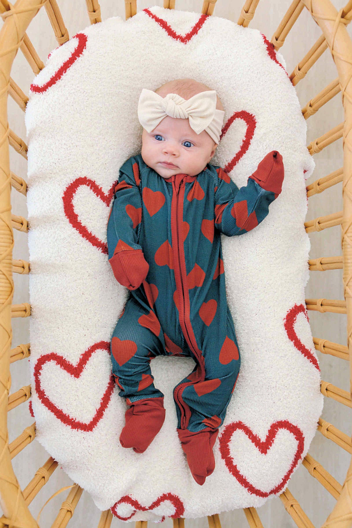 A baby lies on a heart-patterned blanket, wearing a navy bamboo zipper pajama with red hearts and a cream headband.