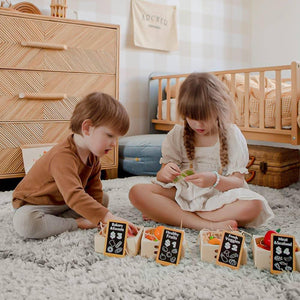 Two children play on a soft, textured rug in a cozy room, examining colorful food items in small wooden crates labeled with different food categories.
