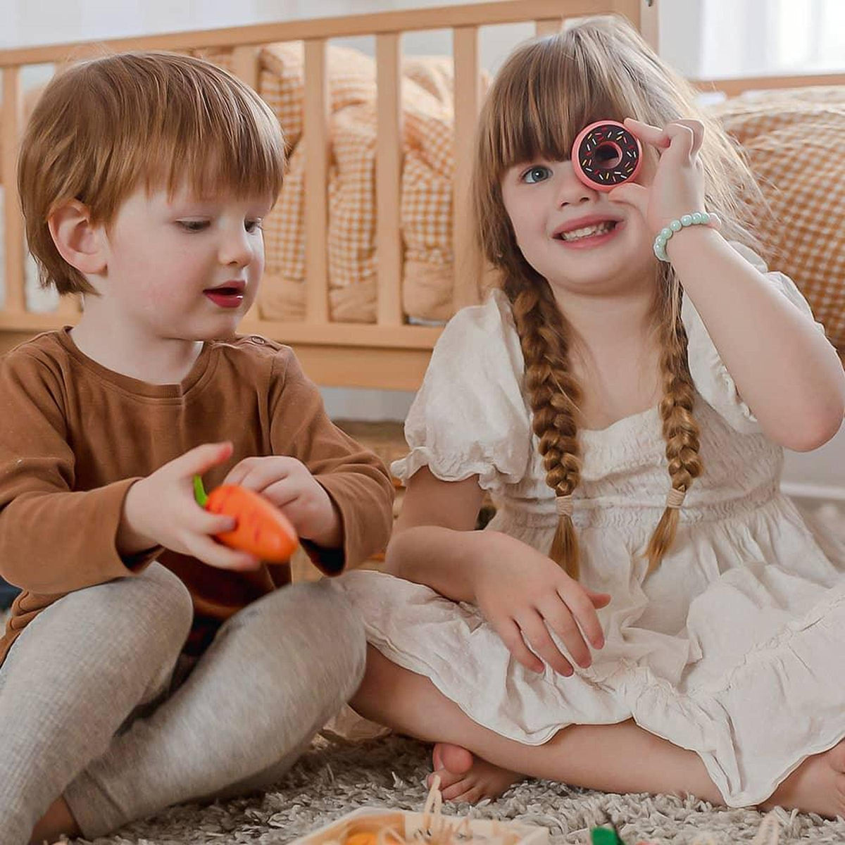 Two children play on a soft carpet, with one girl holding a toy donut and smiling, while the boy beside her holds a toy carrot.
