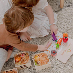 Two children playing on a soft rug, engaged with colorful wooden play food and shopping accessories. They use a toy hammer to interact with a market-themed activity sheet.