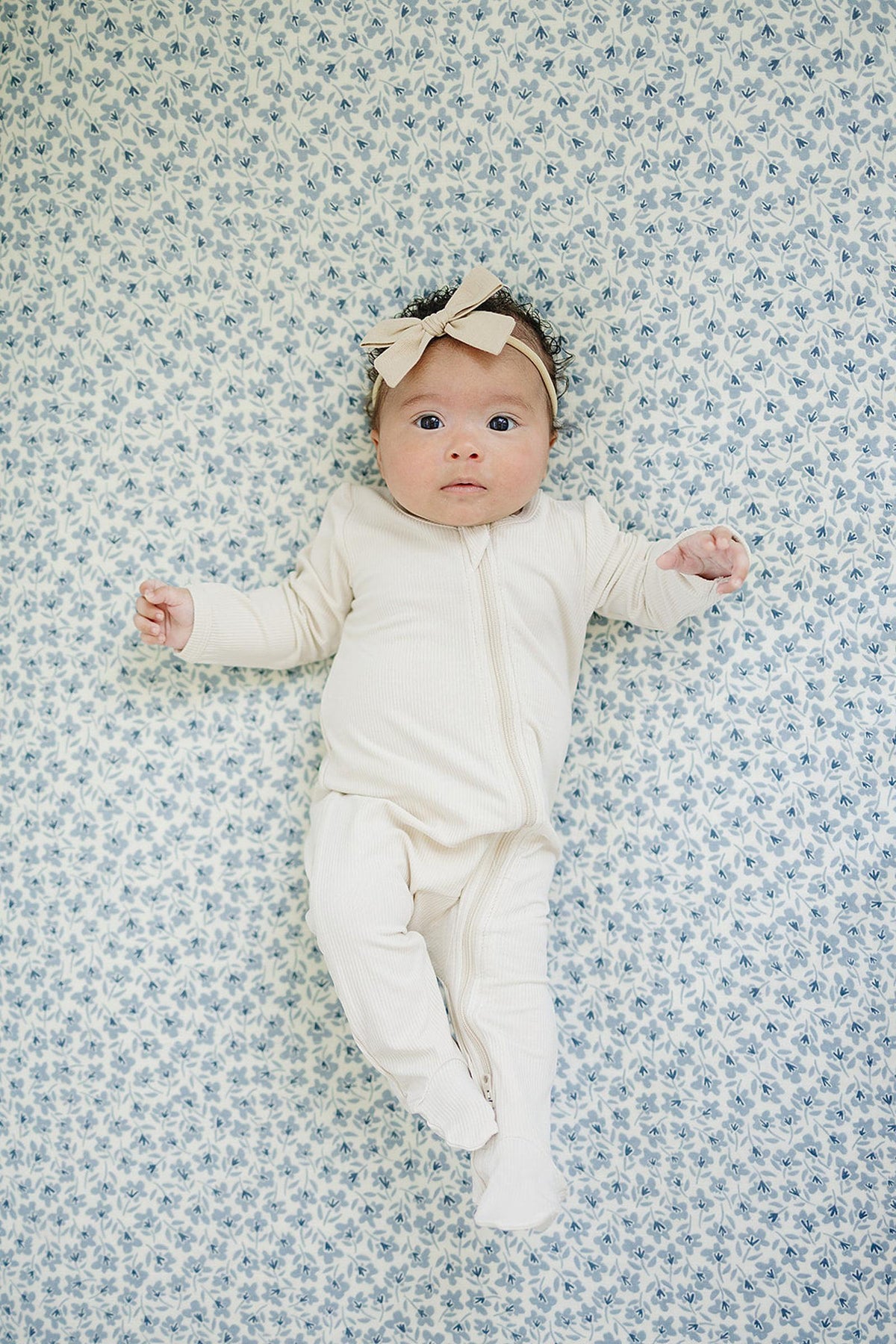 A baby in a cream onesie and bow headband lies on a blue floral-patterned crib sheet.