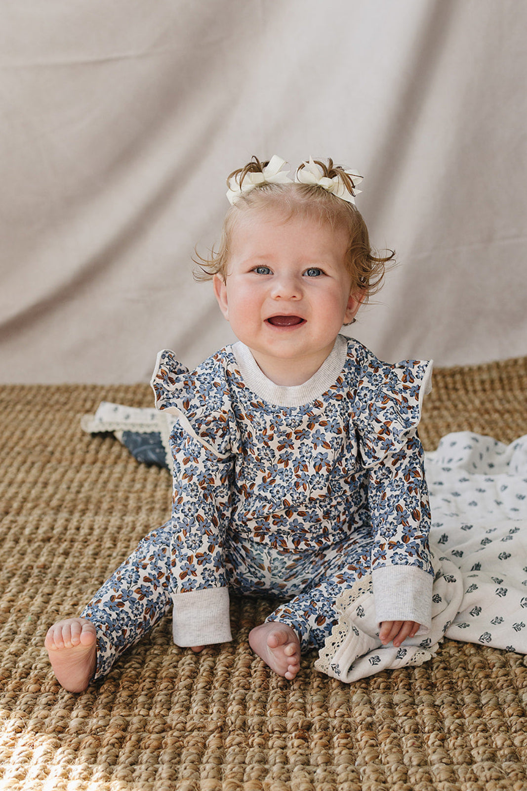 A joyful baby sits on a textured rug, wearing a floral long sleeve cotton outfit with ruffle details. The soft, organic fabric offers comfort, ideal for everyday adventures.