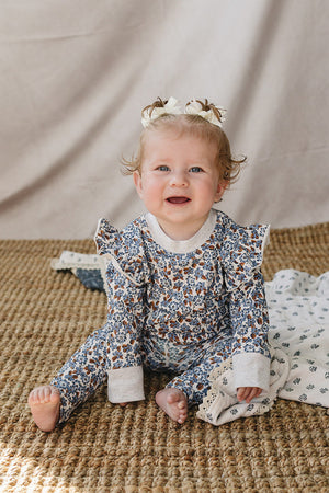 A joyful baby sits on a textured rug, wearing a floral long sleeve cotton outfit with ruffle details. The soft, organic fabric offers comfort, ideal for everyday adventures.