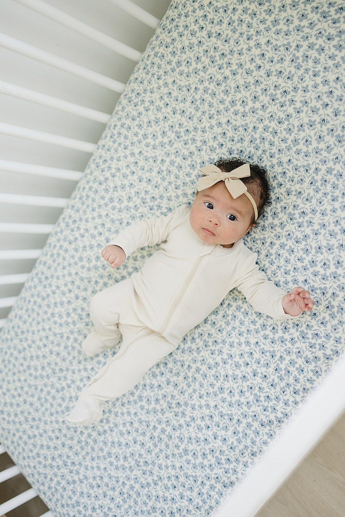 A baby in a beige outfit and headband lies on a blue floral muslin crib sheet in a white crib.