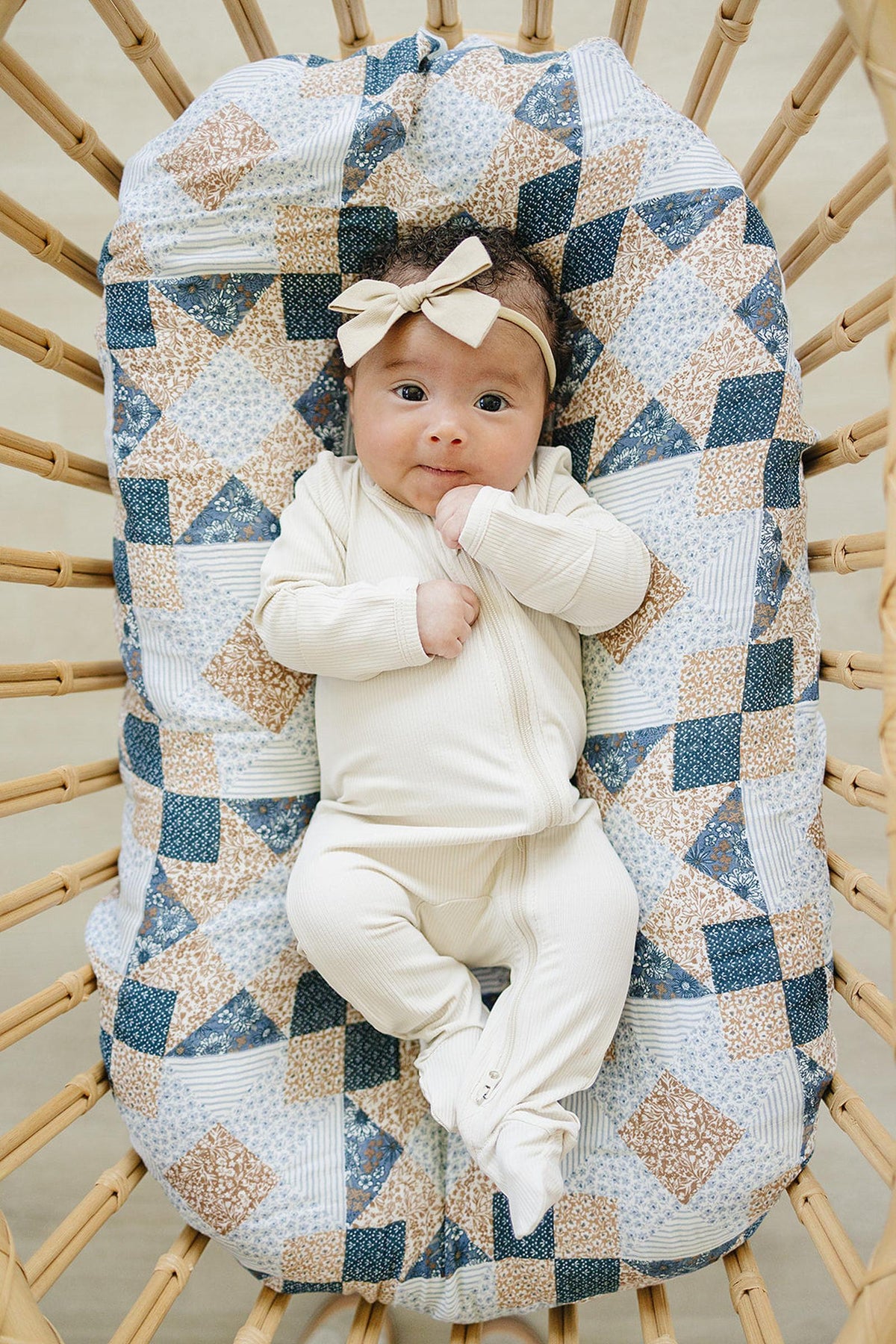 A baby in a cream outfit lies on a patchwork print muslin blanket in a woven crib.