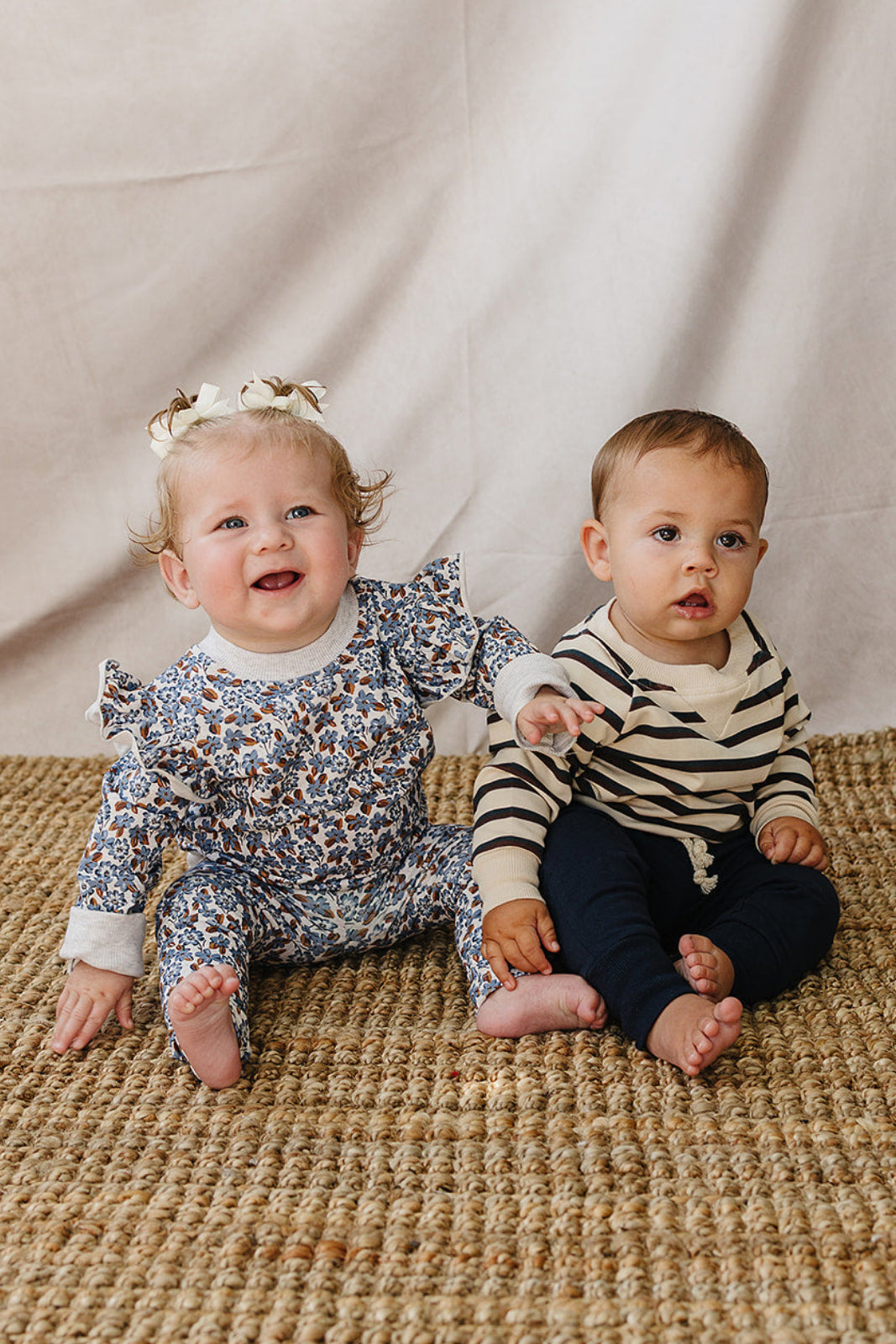 Two toddlers sit on a textured, natural woven rug. One wears a floral-patterned ruffle outfit, while the other is dressed in a striped long-sleeve shirt and navy pants.