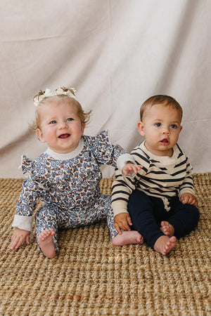 Two toddlers sit on a woven rug, one wearing a floral-patterned outfit with ruffles and the other in a striped crewneck sweatshirt. Both children are smiling, creating a cheerful scene.