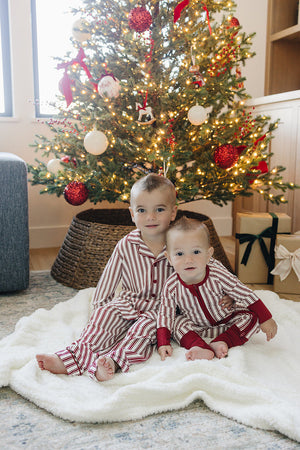 Two children sit on a cozy white blanket in front of a beautifully decorated Christmas tree, wearing matching red-striped bamboo pajama sets. The scene radiates holiday warmth and joy.