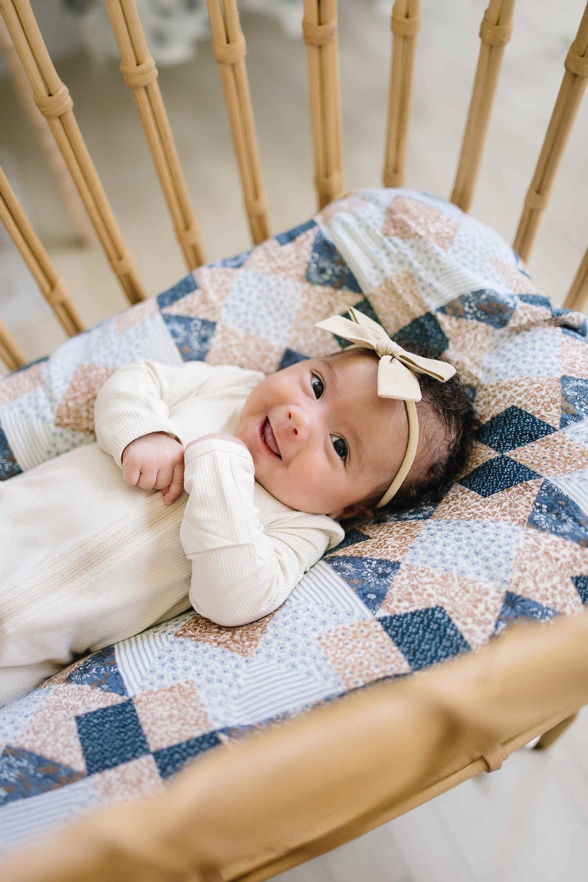 A smiling baby lies on a patchwork print muslin swaddle blanket in neutral colors, wearing a cream headband.