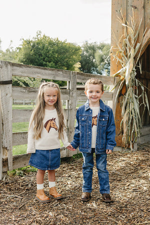 Two children stand hand in hand in a rustic outdoor setting. The girl wears a horse-printed sweater and denim skirt, while the boy sports a denim jacket and matching jeans.