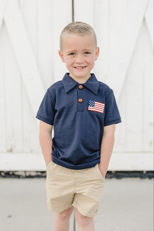 A young boy stands smiling in a classic navy polo featuring a small American flag patch. He pairs it with light khaki shorts, set against a white wooden backdrop.