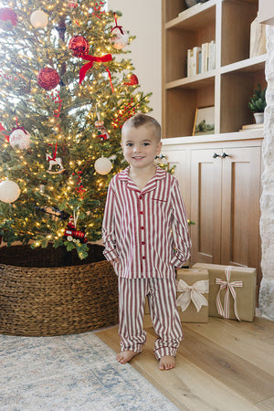 A young child stands barefoot in front of a decorated Christmas tree, wearing a red and white striped collared pajama set. The cozy setting features holiday decorations and wrapped gifts.