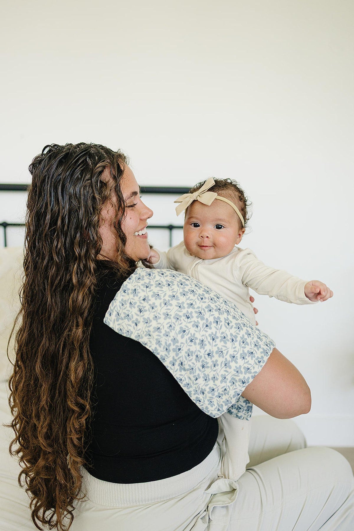 A woman holds a smiling baby wearing a beige headband, with a blue blossom muslin burp cloth draped over her.