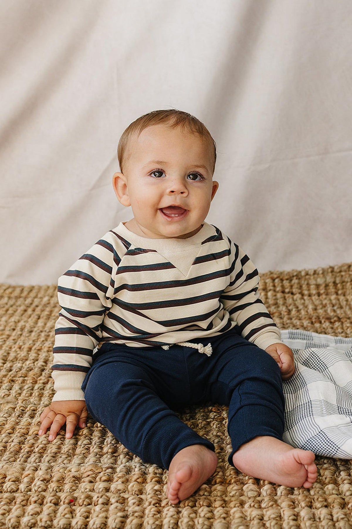 A happy baby sits on a woven rug, wearing a striped cotton crewneck sweatshirt in cream and navy, paired with navy pants. The background features a soft, neutral fabric.