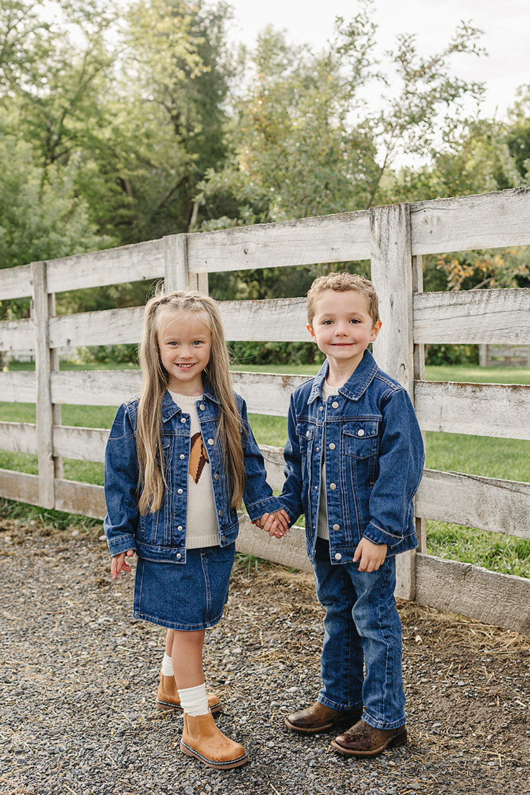 Two children, a girl and a boy, wear matching dark wash denim jackets and jeans, standing hand in hand in front of a rustic wooden fence amid green trees.