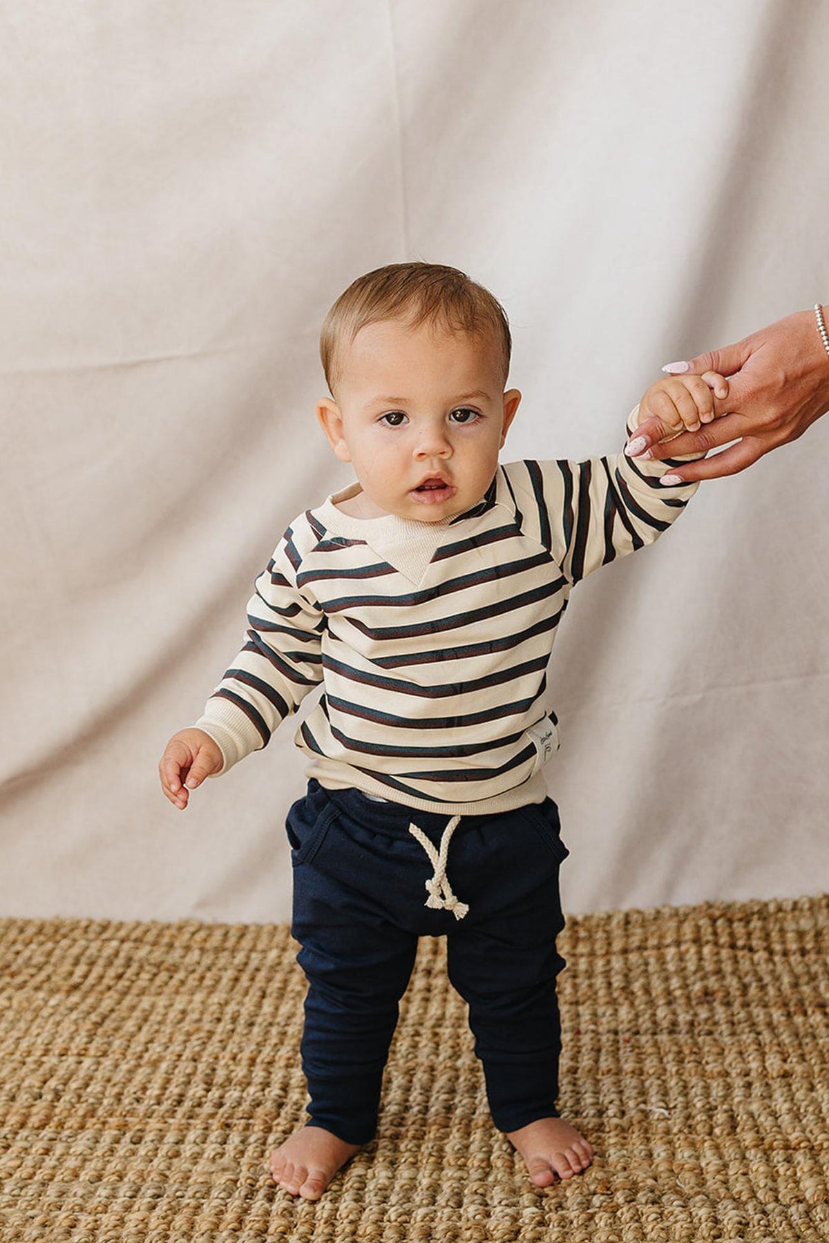 A toddler stands on a woven rug, wearing a striped crewneck sweatshirt in cream and navy. They hold an adults hand, showcasing a casual, playful look.