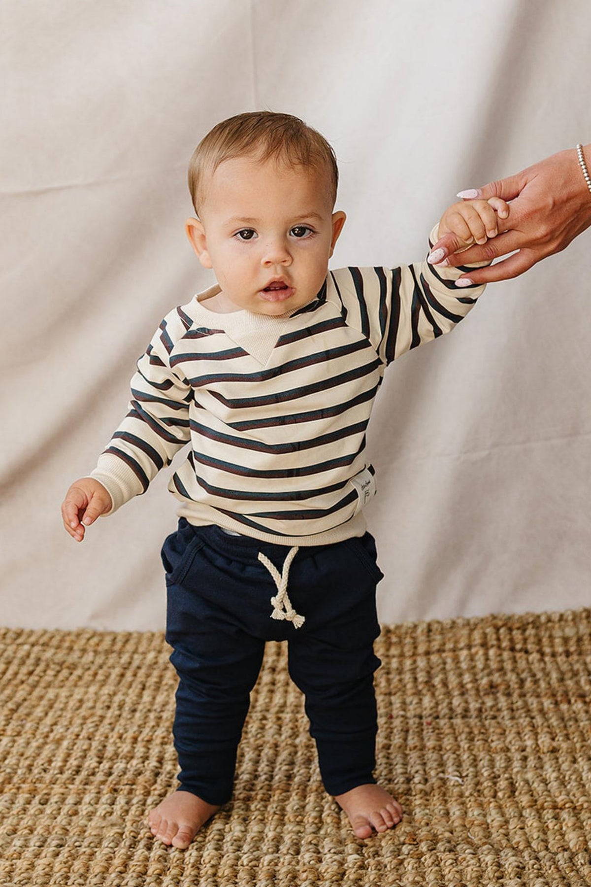 A toddler stands on a textured floor, wearing a striped long-sleeve shirt and navy joggers with a drawstring. An adult gently holds the childs hand, creating a playful atmosphere.