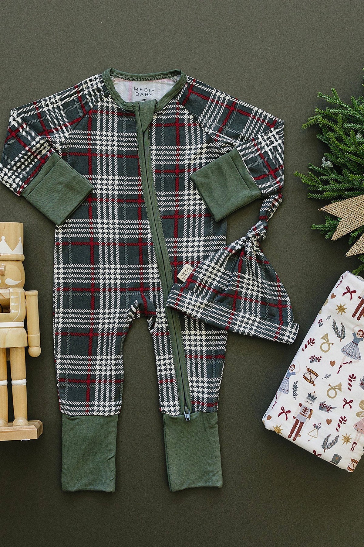 A green plaid bamboo zipper pajama set for babies, featuring long sleeves and matching hat, is displayed on a dark background alongside wooden toys and festive wrapping paper.