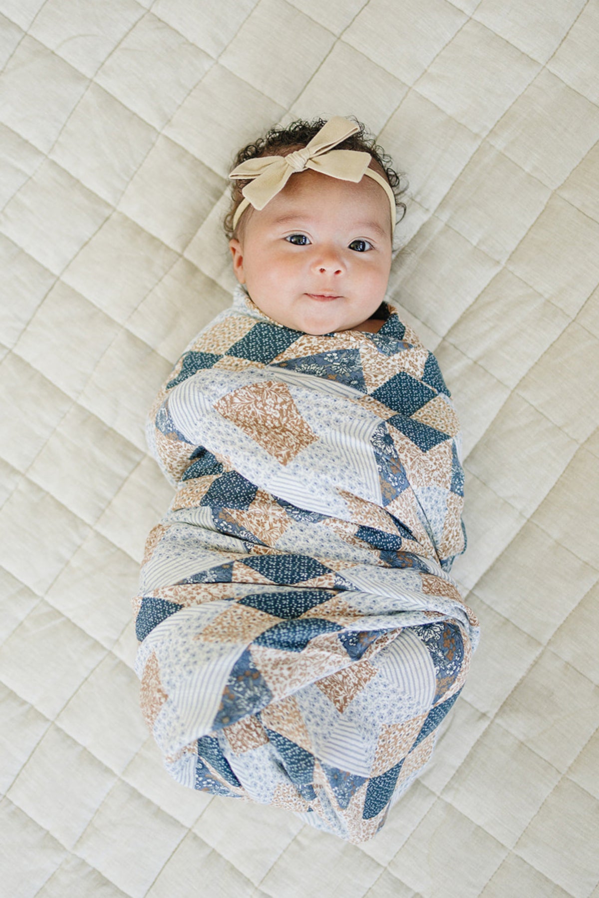 A baby wrapped in a colorful patchwork print muslin swaddle blanket lies on a quilted beige surface.