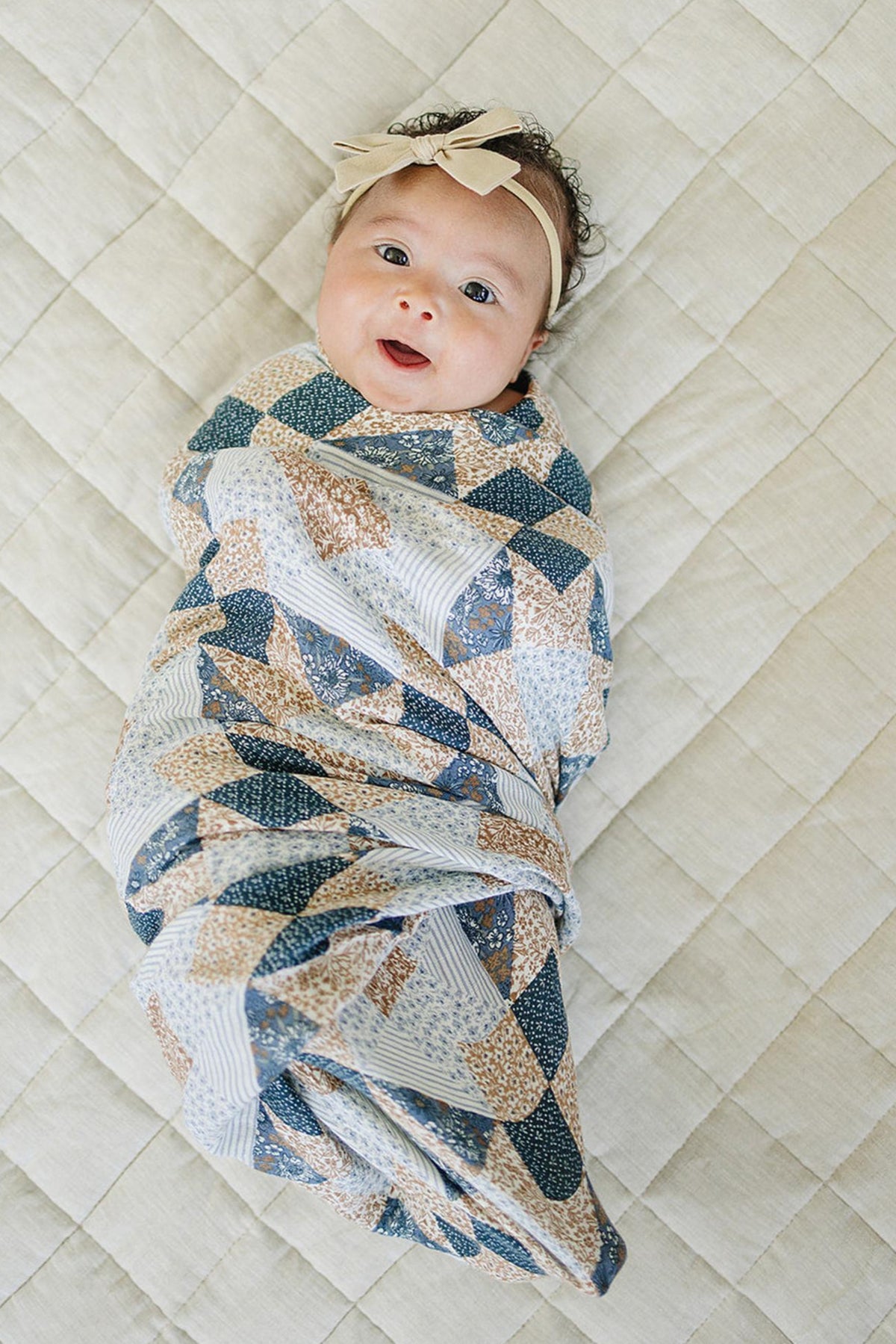 A baby wrapped in a patchwork print muslin swaddle blanket lies on a quilted beige surface.