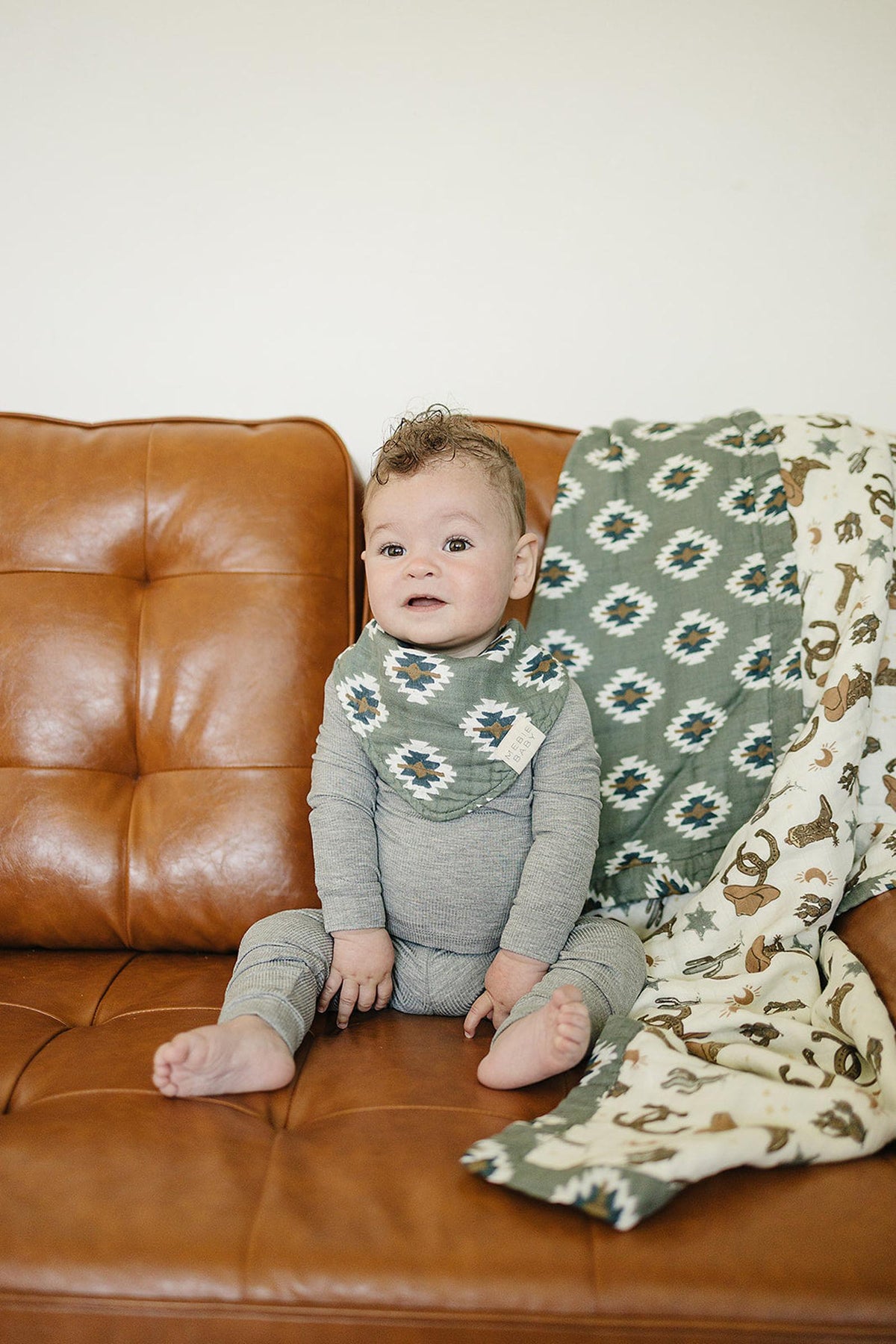 A baby wearing a Southwest Aztec patterned muslin bib sits on a brown leather couch with matching blankets.