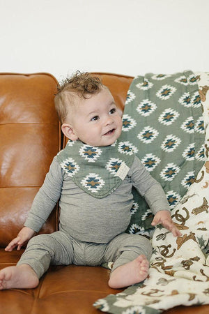 Baby wearing a green Aztec-patterned muslin bib, sitting on a brown leather sofa with matching blanket.