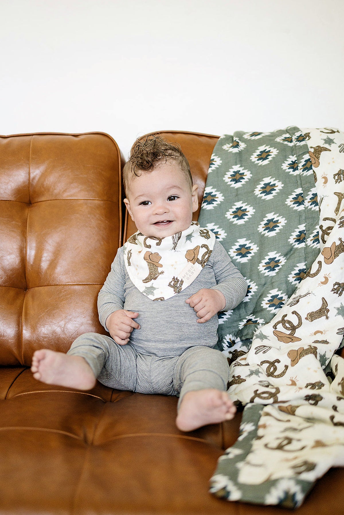 Baby in grey outfit sits on a brown leather couch with a Southwest Aztec quilt and cowboy-themed bib.
