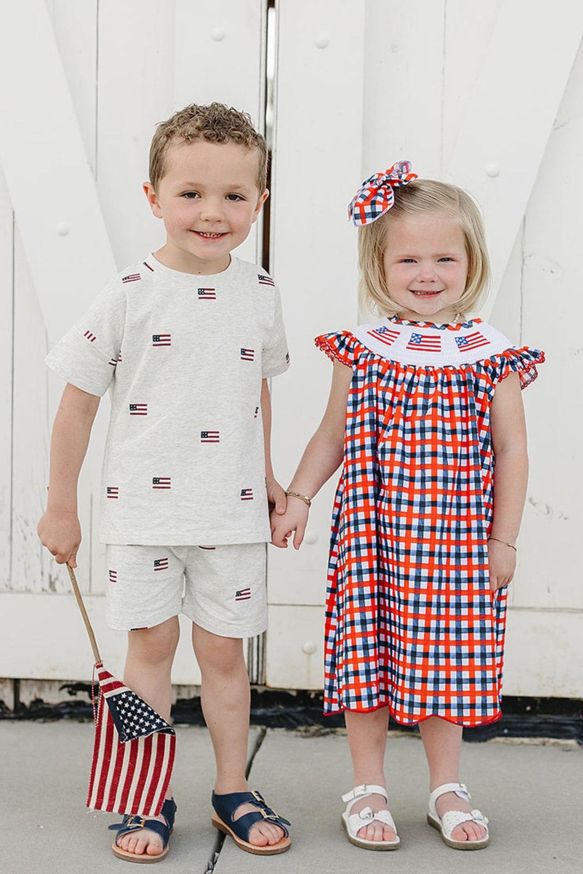 A boy and girl stand together, holding hands in front of a white barn door. The girl wears a red and blue plaid dress with an embroidered collar, while the.