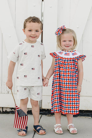 A boy and girl stand together, holding hands in front of a white barn door. The girl wears a red and blue plaid dress with an embroidered collar, while the.