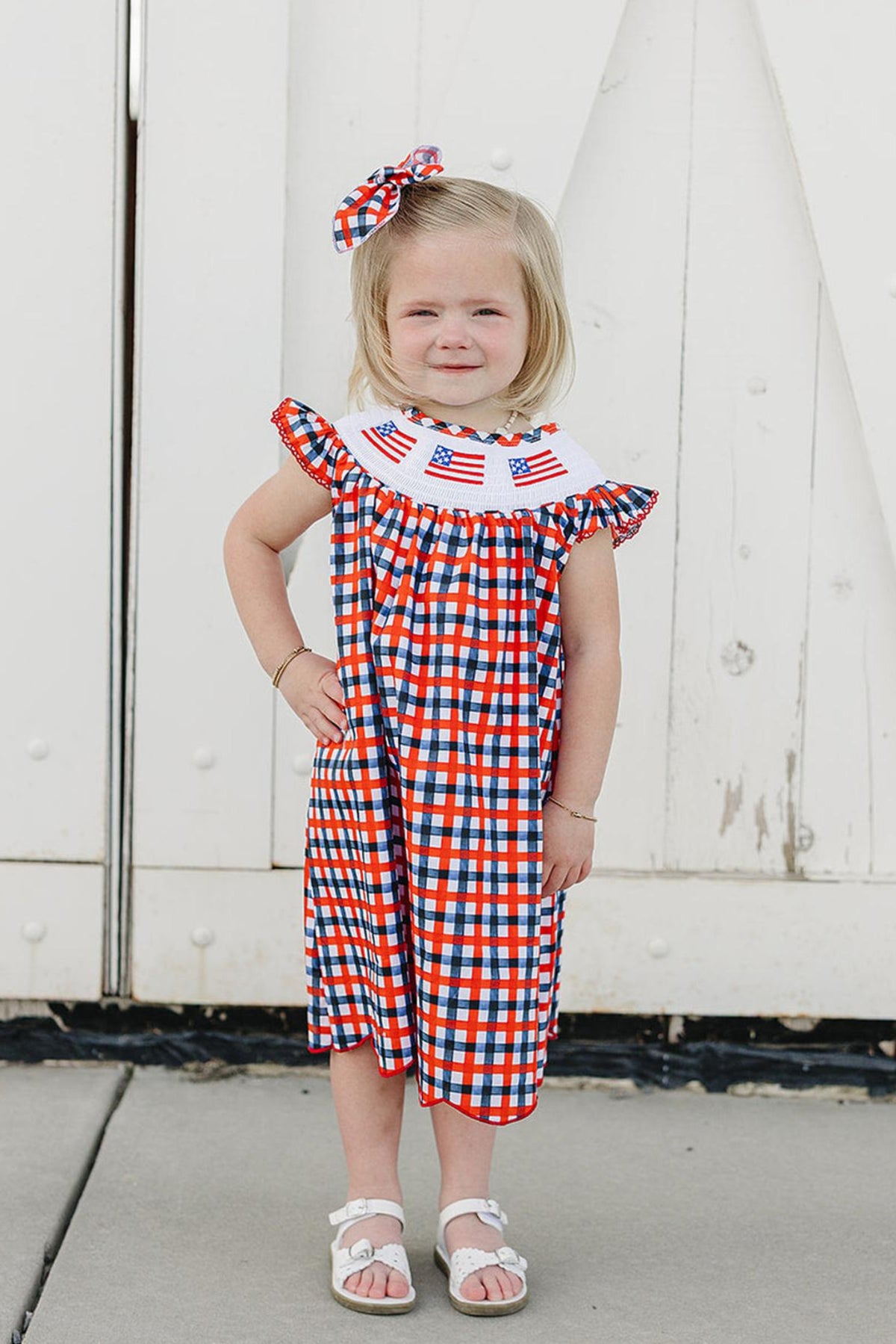 A young girl stands in front of a white barn door, wearing a red and blue gingham dress with a smocked collar featuring embroidered American flags and a matching hair.