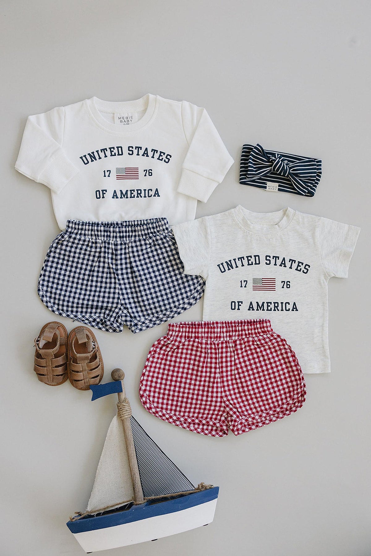 Baby clothes set with United States of America print, navy and red checkered shorts, striped headband, sandals, and toy sailboat.