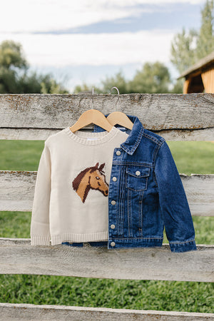 A denim jean jacket hangs on a wooden fence, paired with a cream sweater featuring a horse design. The setting is an open green field under a clear sky.