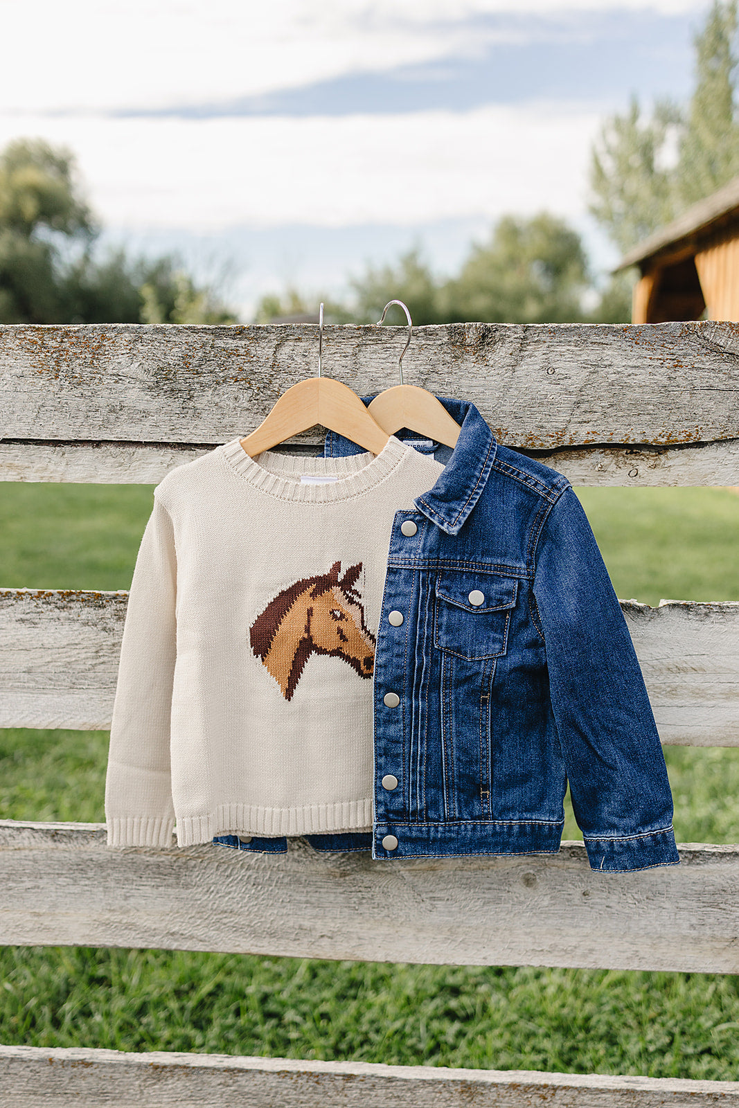 A cozy cream sweater featuring a horse design hangs alongside a denim jacket on a wooden fence. The backdrop showcases a vibrant green field and a clear sky.