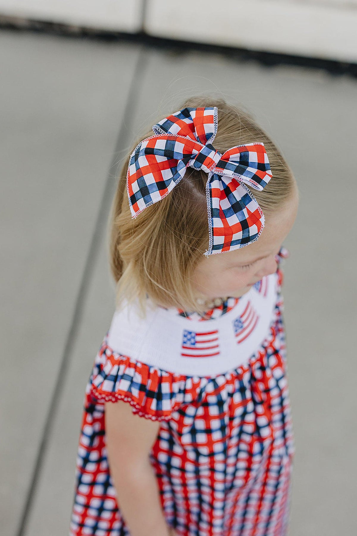 A young girl with a plaid hair bow in red, white, and blue stands outdoors, wearing a matching checkered dress featuring American flags.