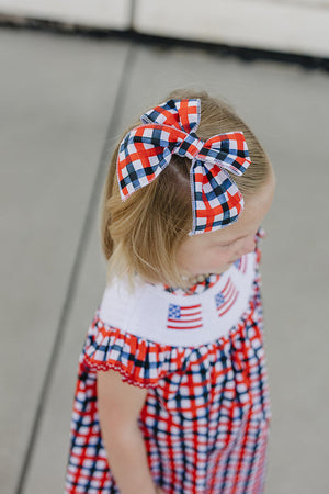 A young girl with a plaid hair bow in red, white, and blue stands outdoors, wearing a matching checkered dress featuring American flags.