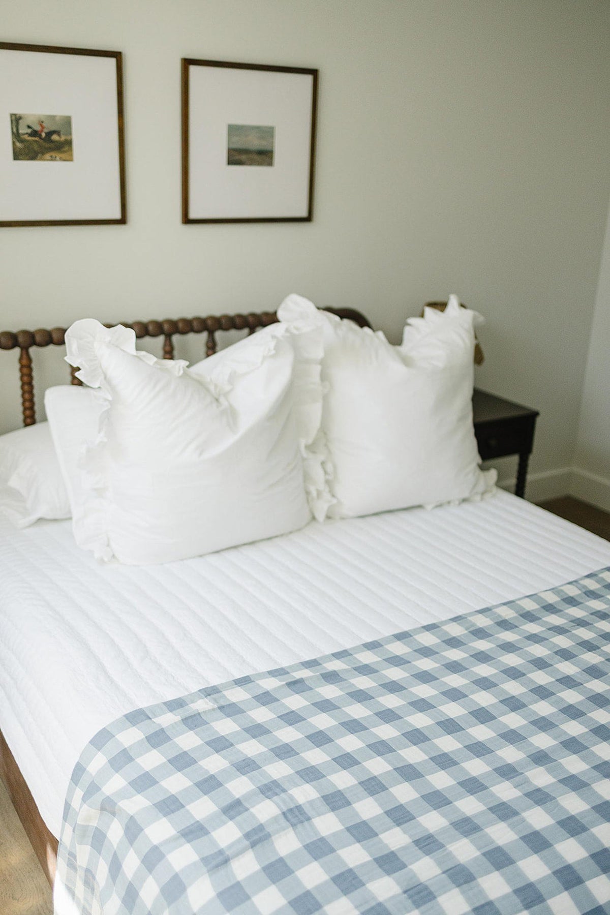 A bedroom features a bed with a dusty blue gingham quilt and white ruffled pillows, against framed artwork.