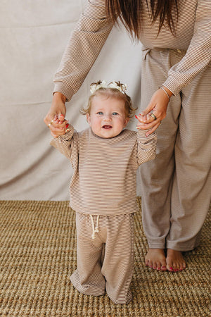 A smiling baby in a soft, striped loungewear set holds hands with an adult. The cozy outfit features a long-sleeve top and pants, perfect for family matching moments.