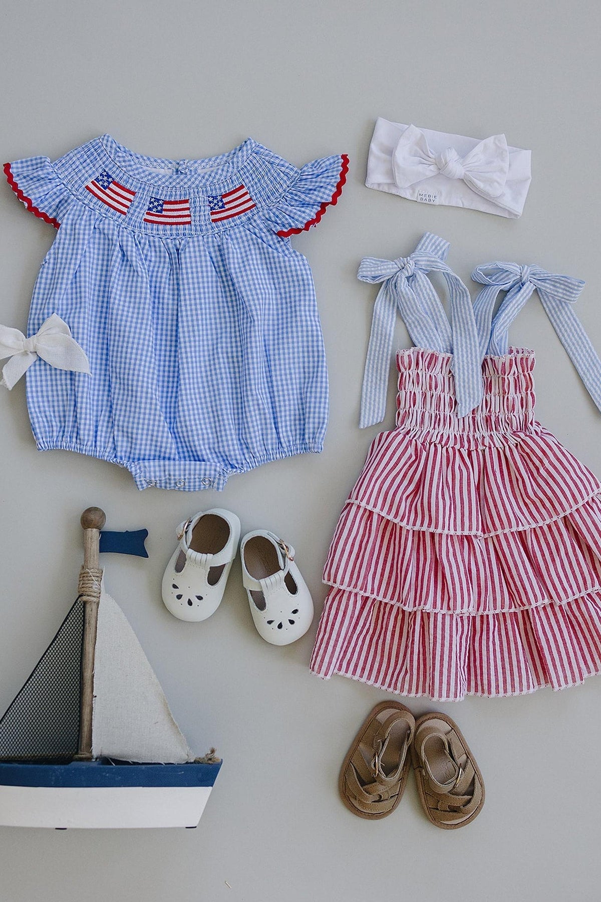 Blue gingham romper with flag embroidery, red striped ruffle dress, headbands, shoes, and a toy sailboat.