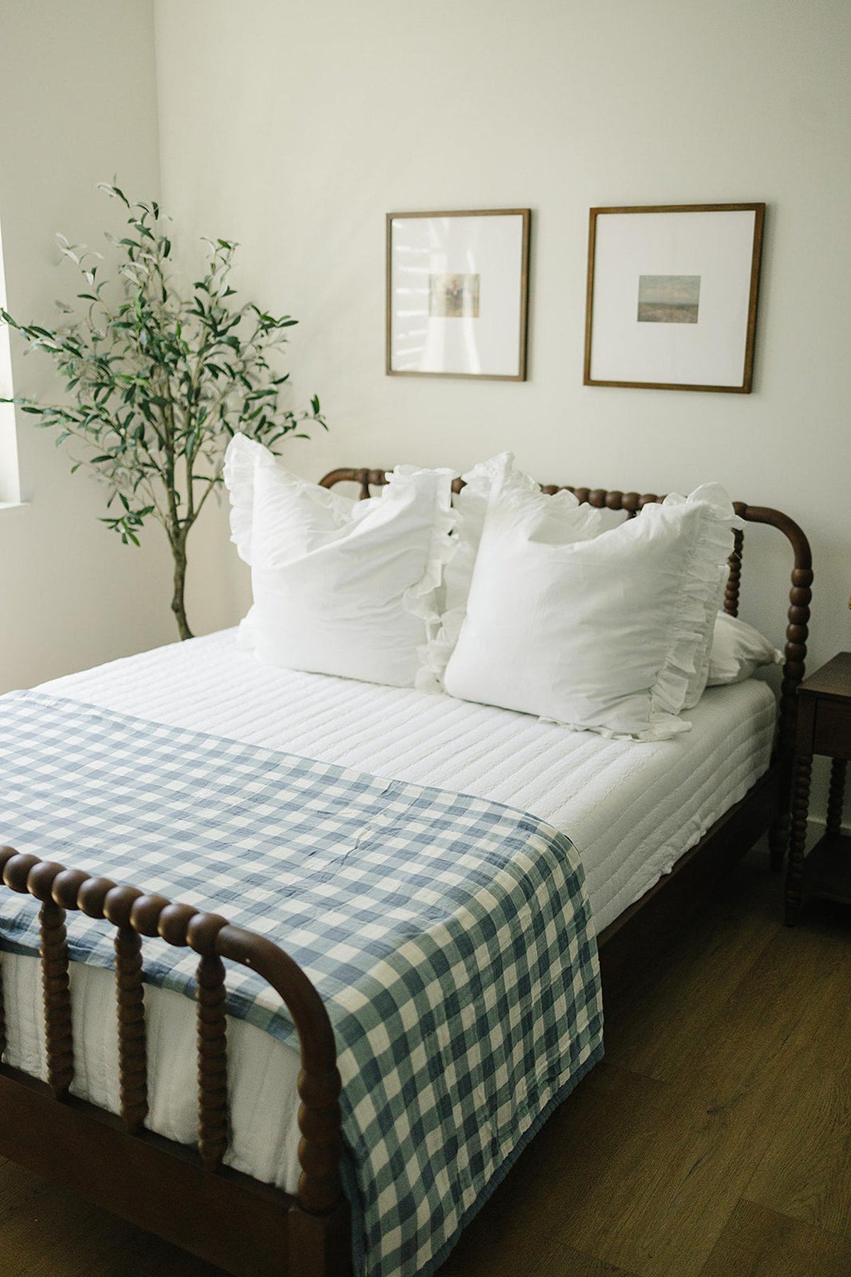 A wooden bed with white pillows and a Dusty Blue Gingham quilt, next to a potted plant.