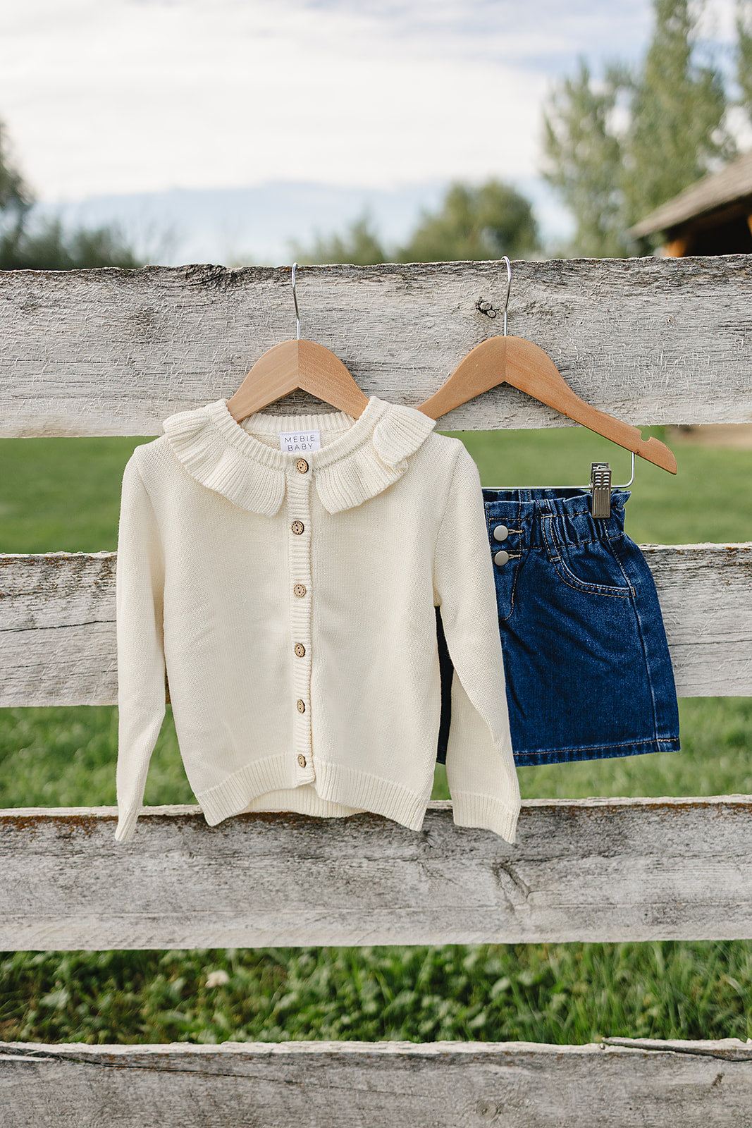 A cream ruffle knit cardigan with a button-down front hangs on a wooden hanger beside a pair of denim shorts against a rustic fence, set in a grassy outdoor environment.