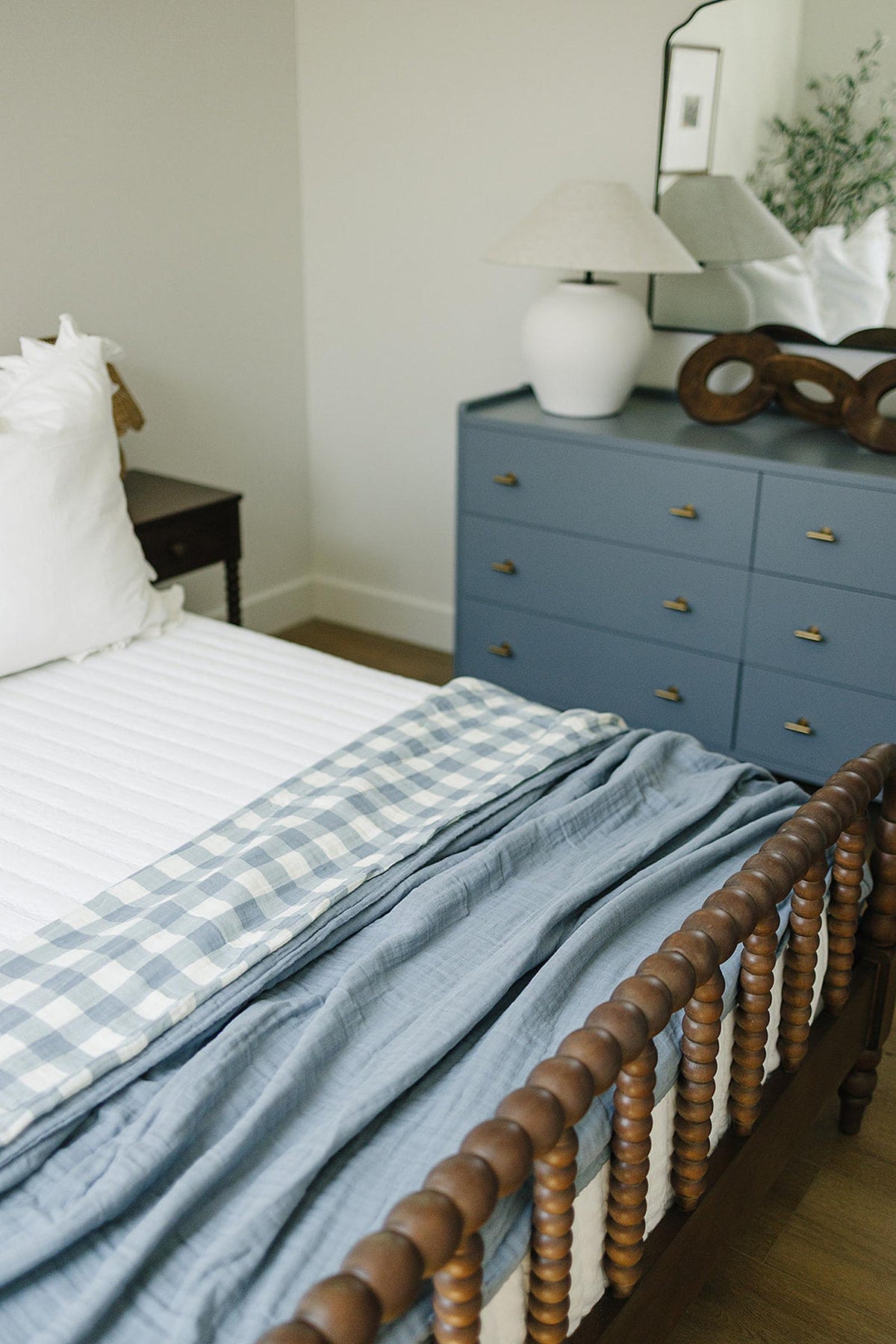 A cozy bedroom with a wooden bed featuring a dusty blue gingham quilt, next to a blue dresser.