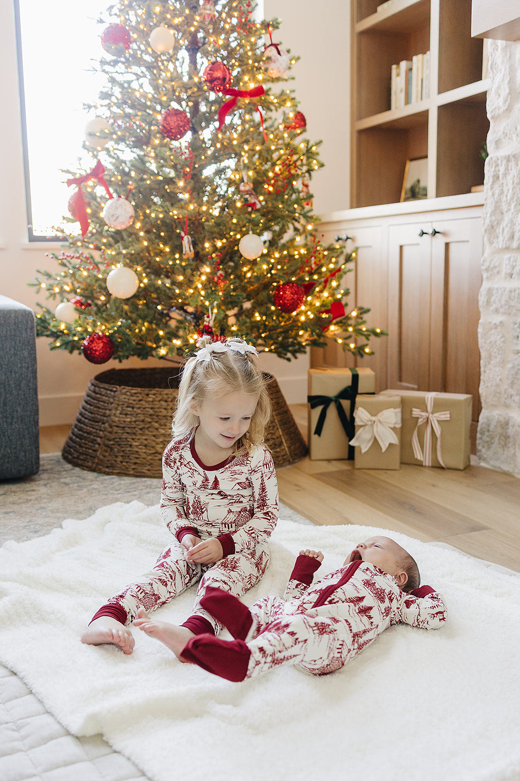 Two children sit on a soft white rug in front of a decorated Christmas tree. They wear a cozy holiday-themed pajama set featuring a classic toile print in festive colors.