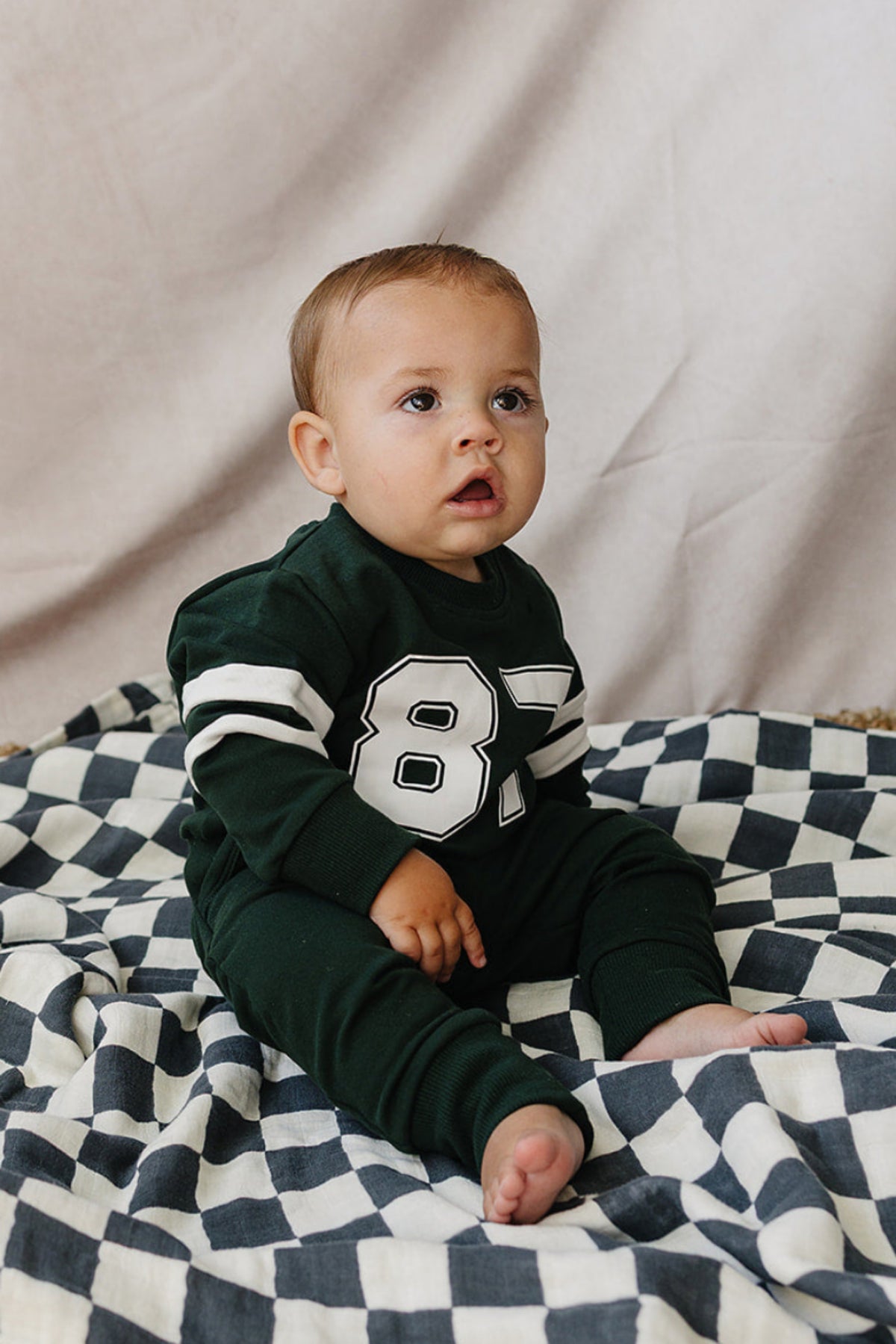 A baby sits on a checkered blanket wearing a dark green French terry cotton set featuring a large number 87 on the front, designed for comfort and everyday adventures.