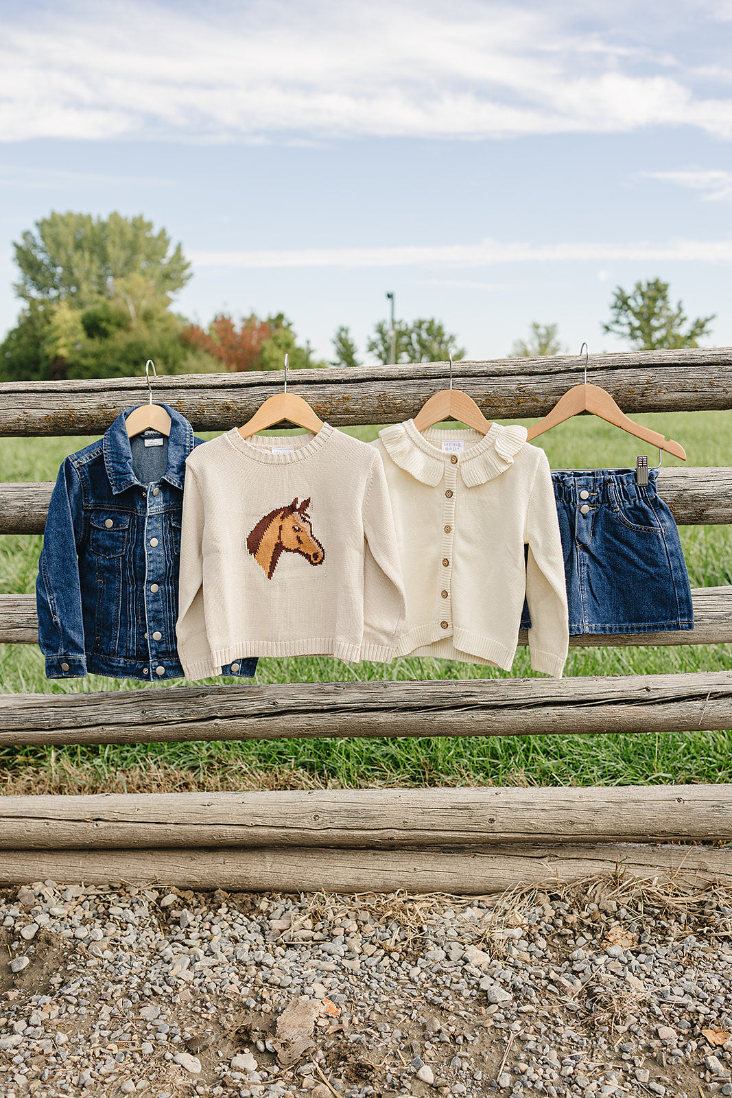 A cozy cream horse knit sweater hangs alongside a denim jacket and a denim skirt against a rustic wooden fence, set against a green field and clear blue sky.