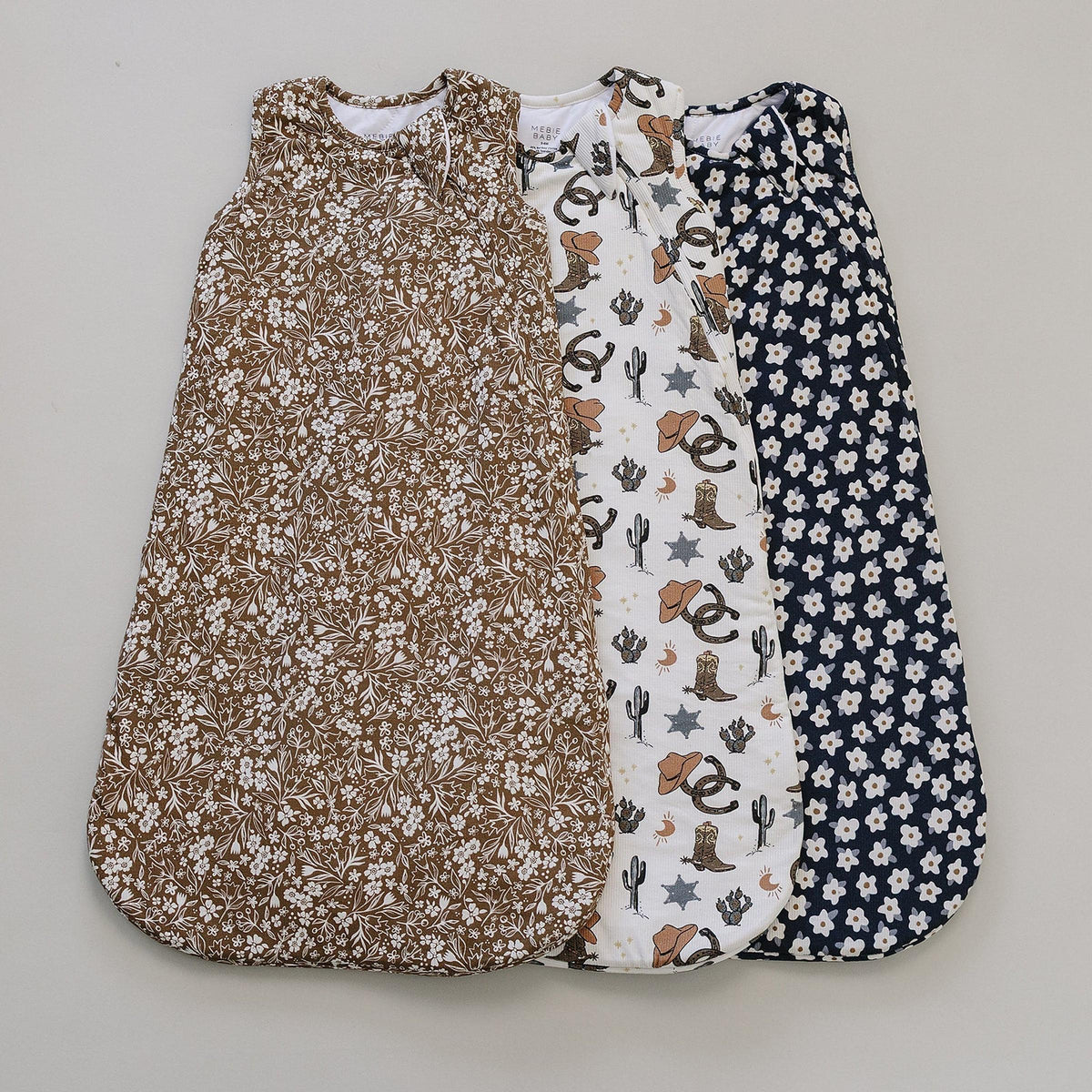 Three beautifully designed baby sleep sacks are displayed, featuring a brown floral pattern, a white print with cacti and animals, and a navy floral design, all made from soft, breathable.