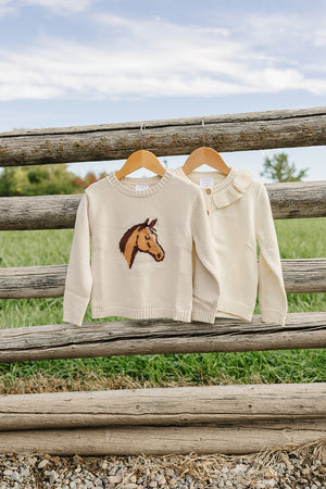 Two cream-colored knit cardigans hang on wooden hangers against a rustic fence, featuring a horse design on one and a ruffled neckline on the other, perfect for childrens cozy outfits.