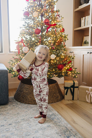 A young child in a festive holiday-themed pajama set stands next to a decorated Christmas tree, holding a gift.
