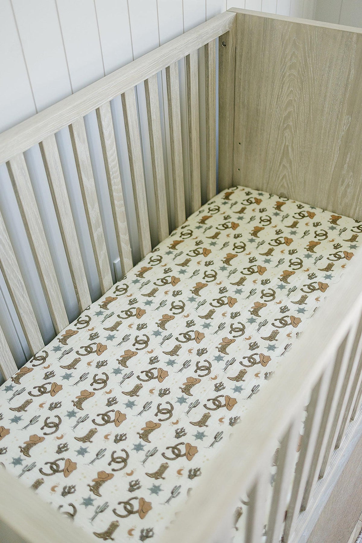 A wooden crib features a muslin crib sheet with cowboy-themed prints, including hats, boots, and cacti.
