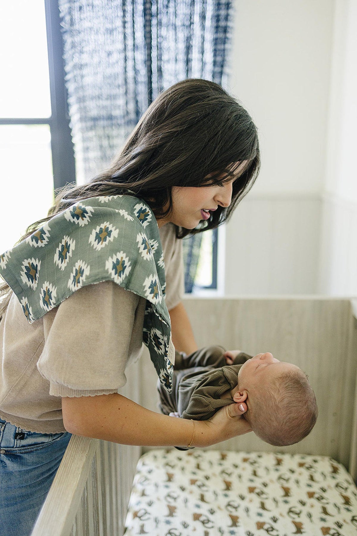 A woman holds a baby in a crib, wearing a Southwest Aztec muslin burp cloth over her shoulder.