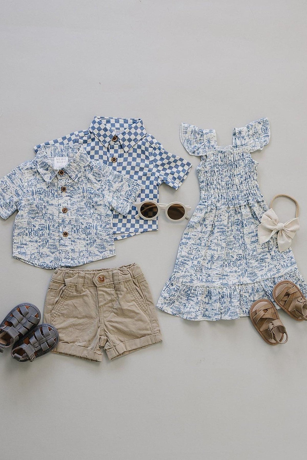 Flat lay of two boys button-up shirts in light blue patterns, beige shorts, and a girls dress with ruffled sleeves, paired with sunglasses and sandals, showcasing a stylish summer outfit.